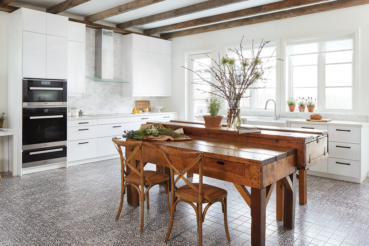 Modern, white farmhouse kitchen with rustic beams, worktables and patterned tile floors.